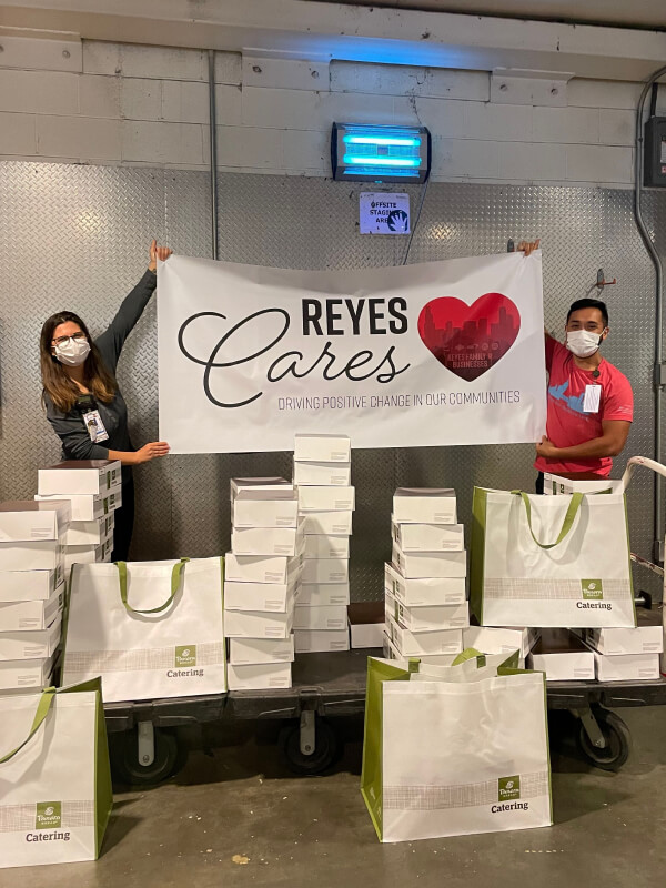 Two employees wearing masks and holding "Reyes Cares" poster surrounded by many Panera Bread Catering bags and boxes