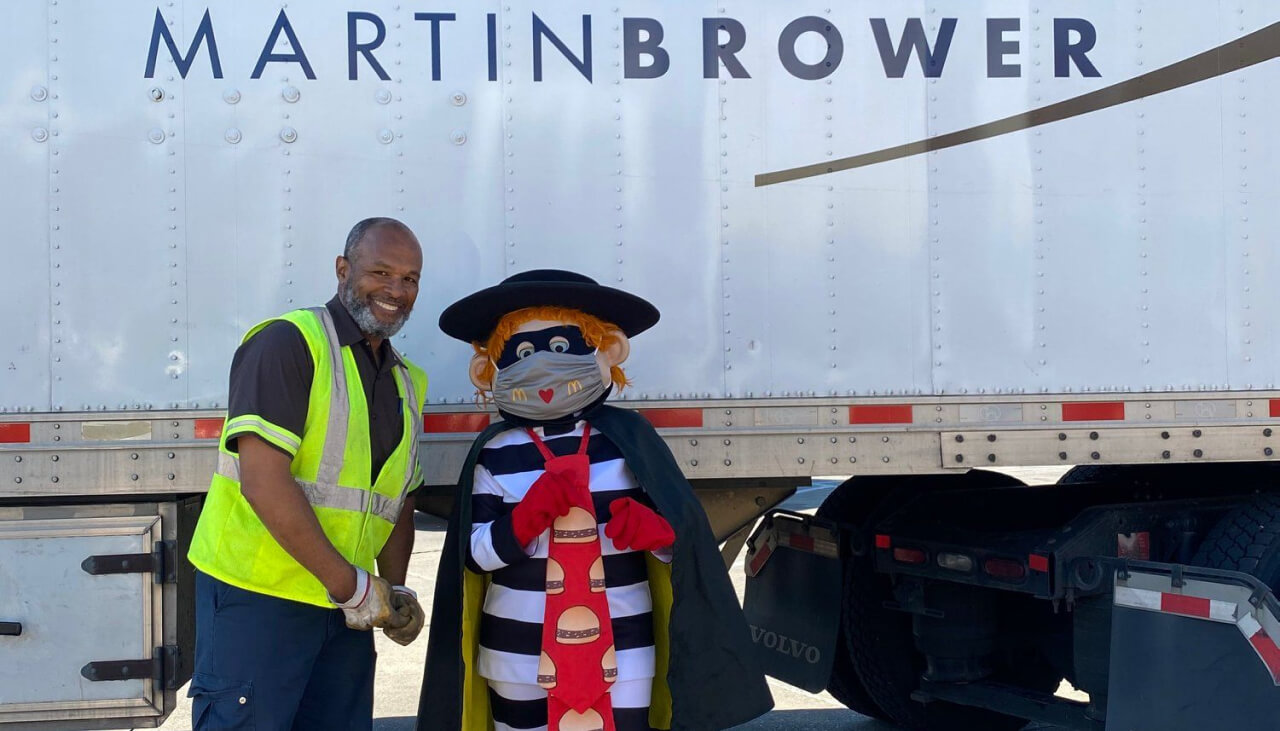 Smiling man standing next to another employee who is wearing Macdonald's Zorro Mask Costume and a tie with burgers on it. Martin Brower truck in the background.