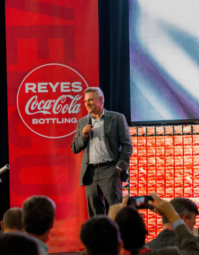 Man wearing a suit and holding a microphone on the stage with Coca-Cola banner and boxes in the background
